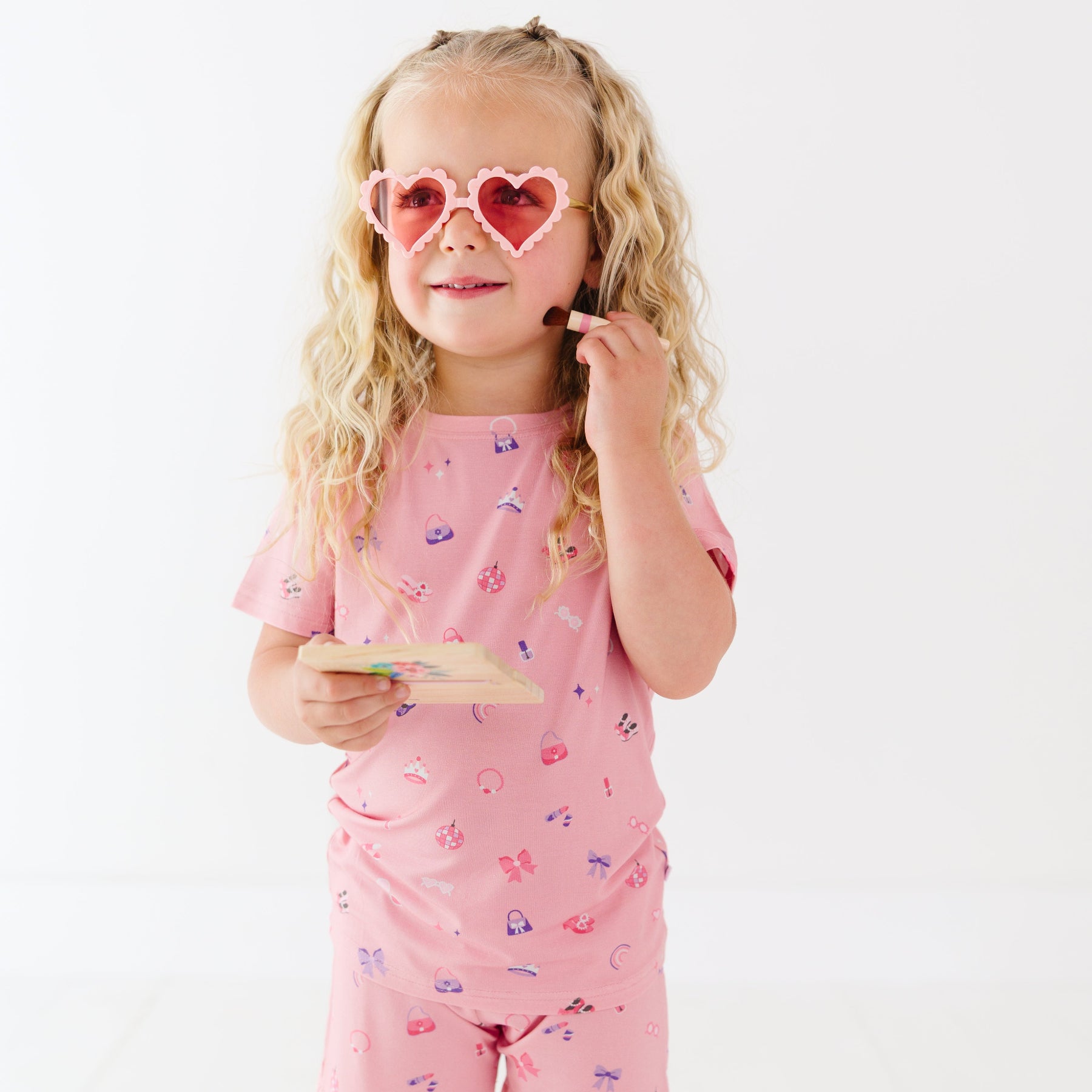 Young girl wearing pink sunglasses with heart shapes, holding a brush and make-up palette on a white background