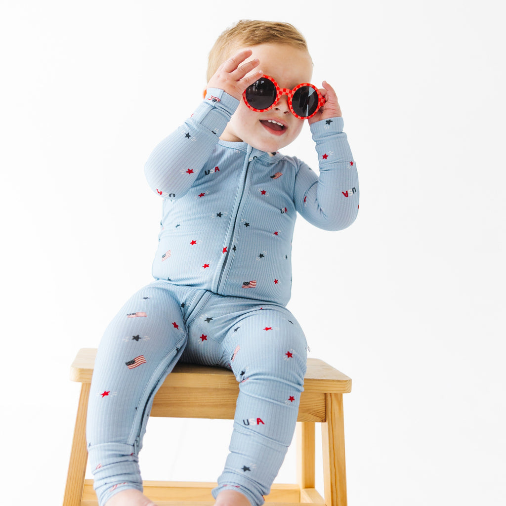 Child wearing a light blue onesie with stars and flag patterns, wearing red sunglasses sitting on a stool.