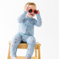 Child wearing a light blue onesie with stars and flag patterns, wearing red sunglasses sitting on a stool.
