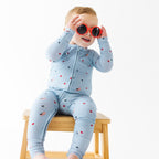 Child wearing a light blue onesie with stars and flag patterns, wearing red sunglasses sitting on a stool.