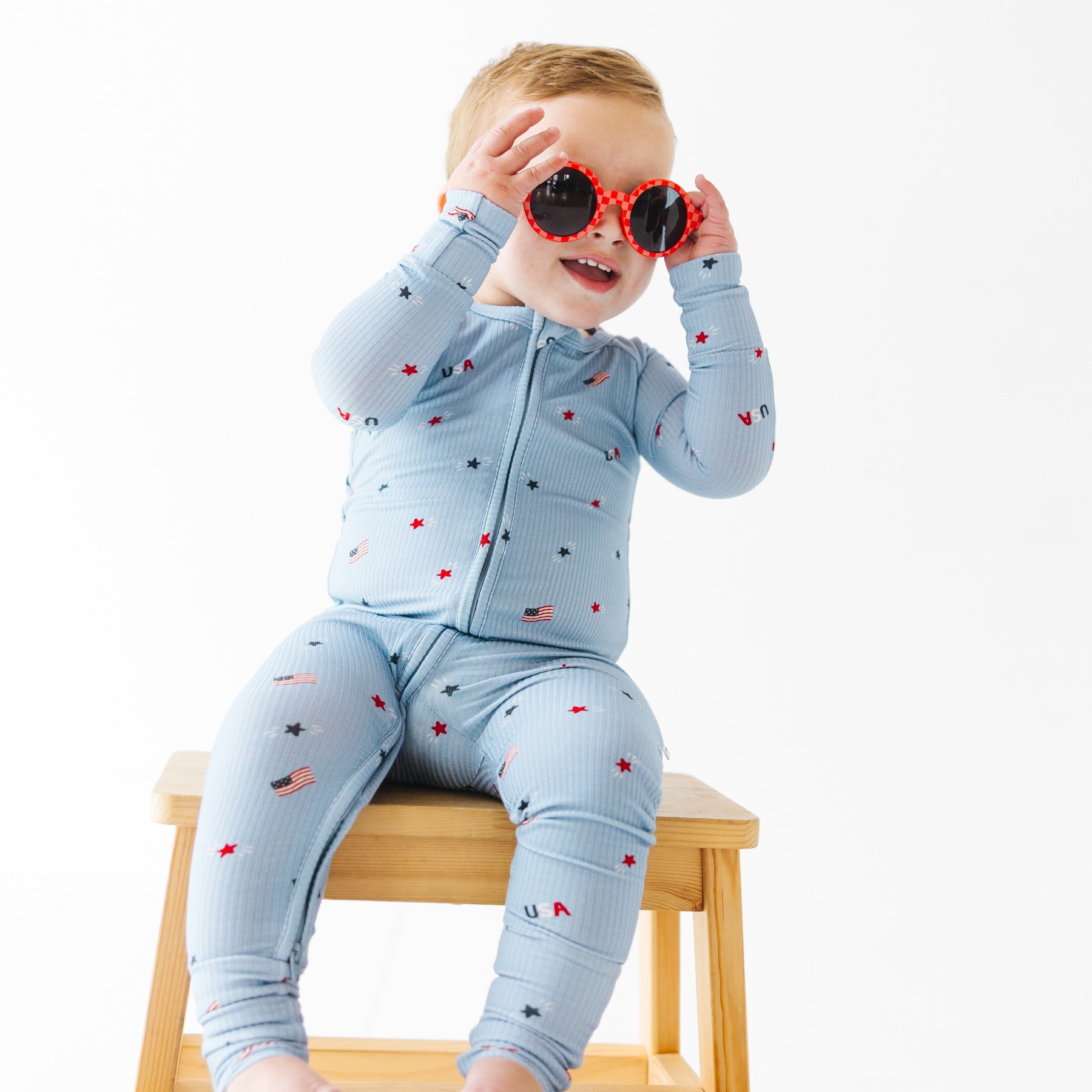 Child wearing a light blue onesie with stars and flag patterns, wearing red sunglasses sitting on a stool.