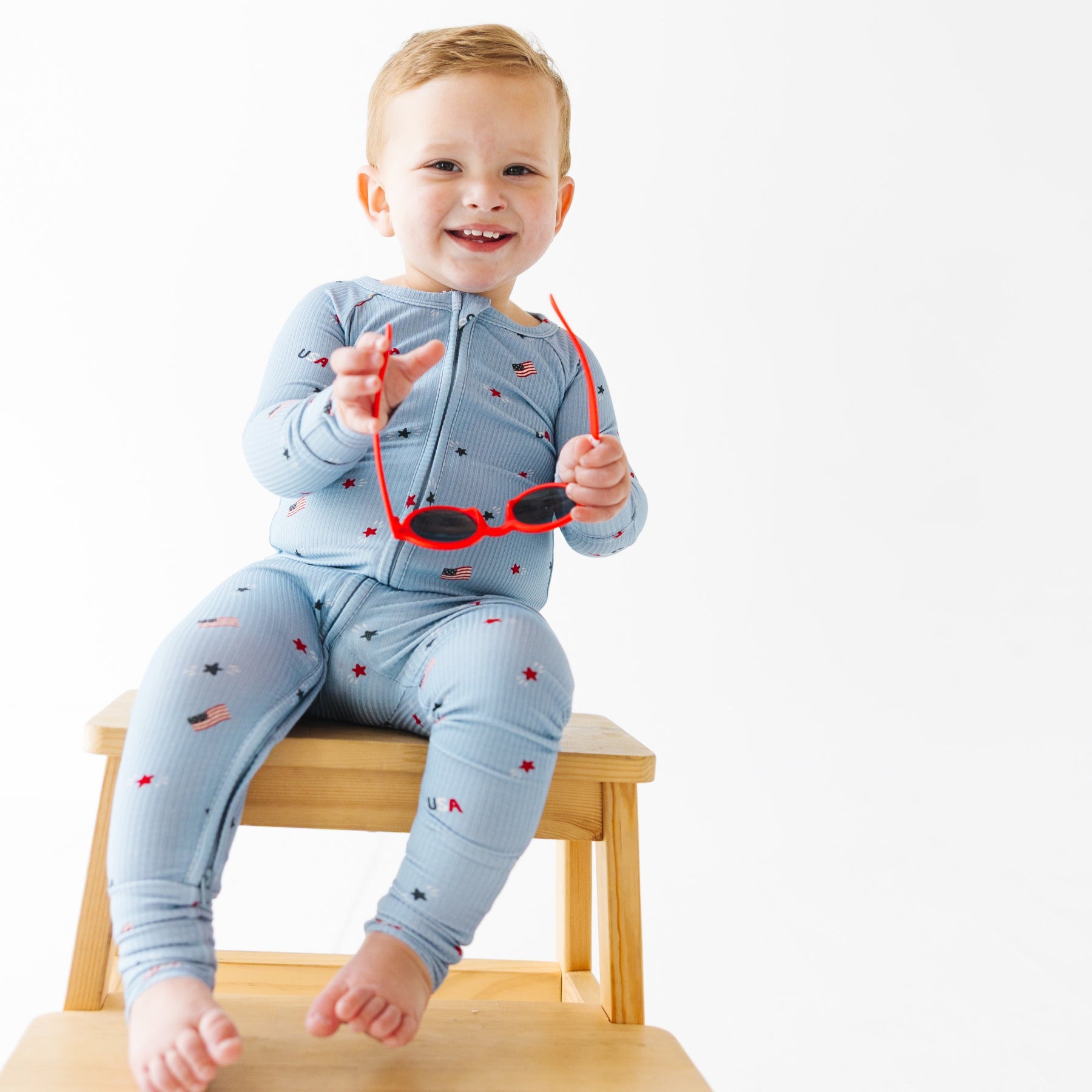 Child wearing a light blue onesie with stars and flag patterns, holding red sunglasses sitting on a stool.