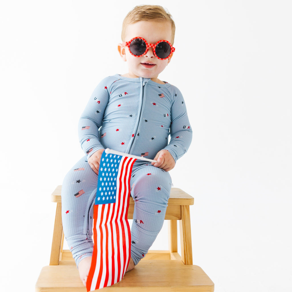 Child wearing a light blue onesie with stars and flag patterns, wearing red sunglasses and holding a USA flag while sitting on a stool.