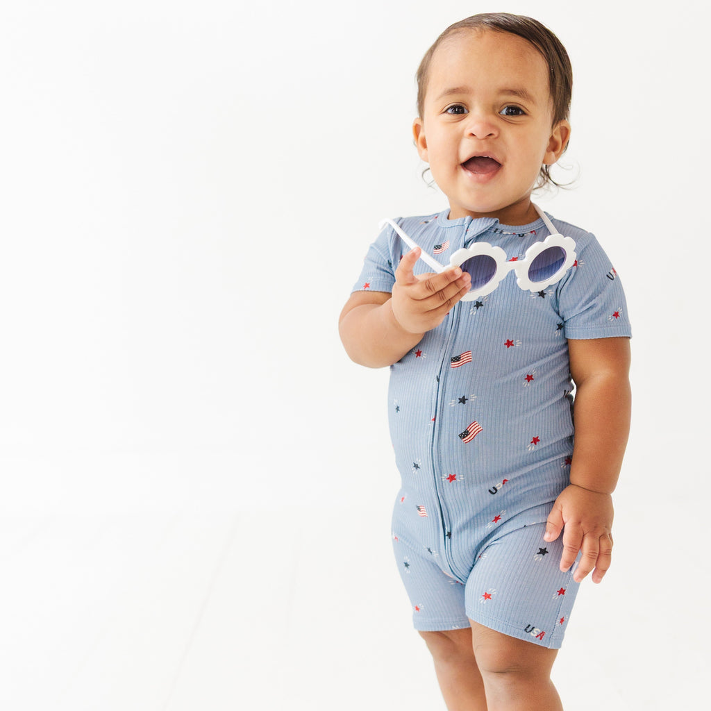 Child in blue short romper with star pattern, holding sunglasses on a white background