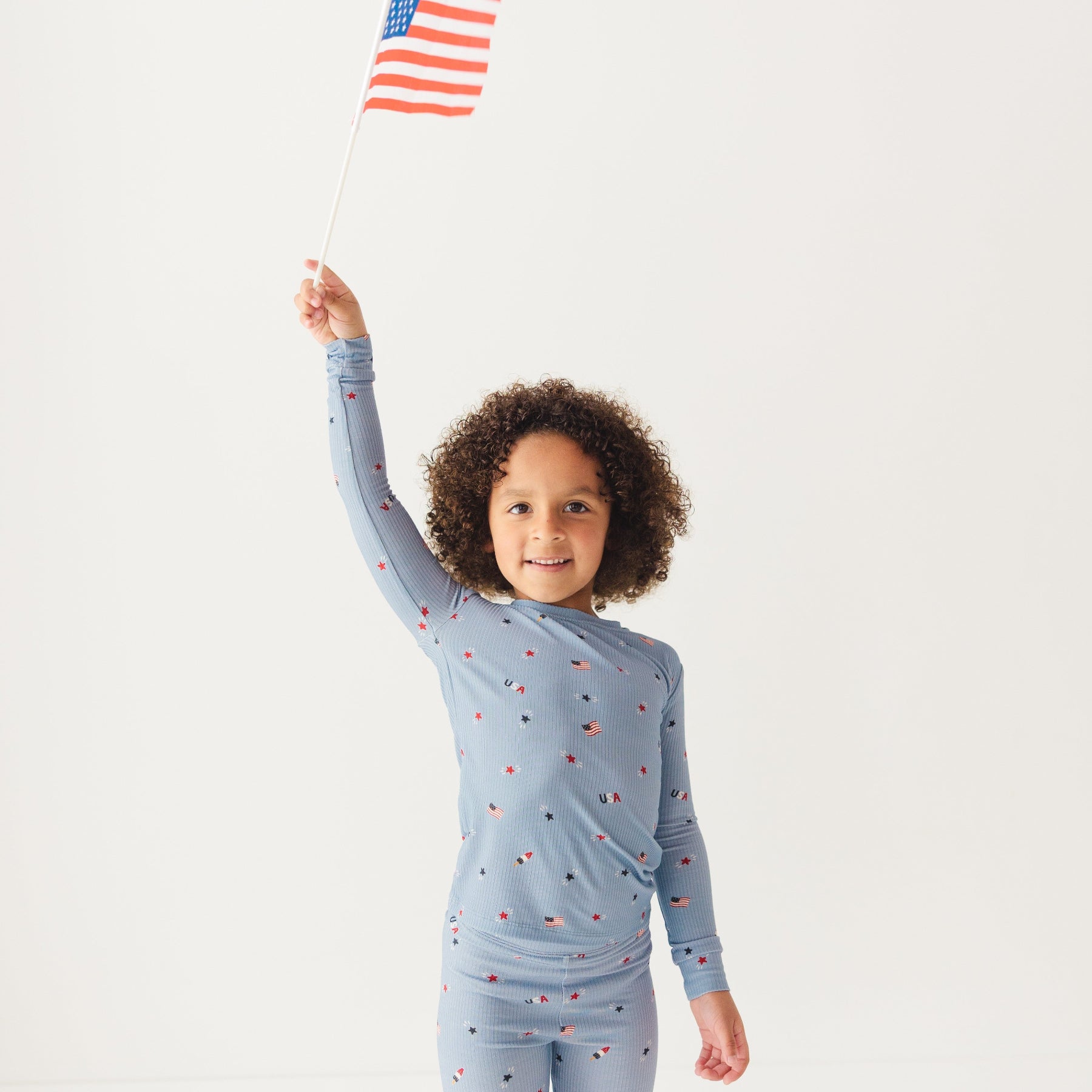 Child wearing a blue pajama set with stars and flag patterns, holding an American flag on a white background