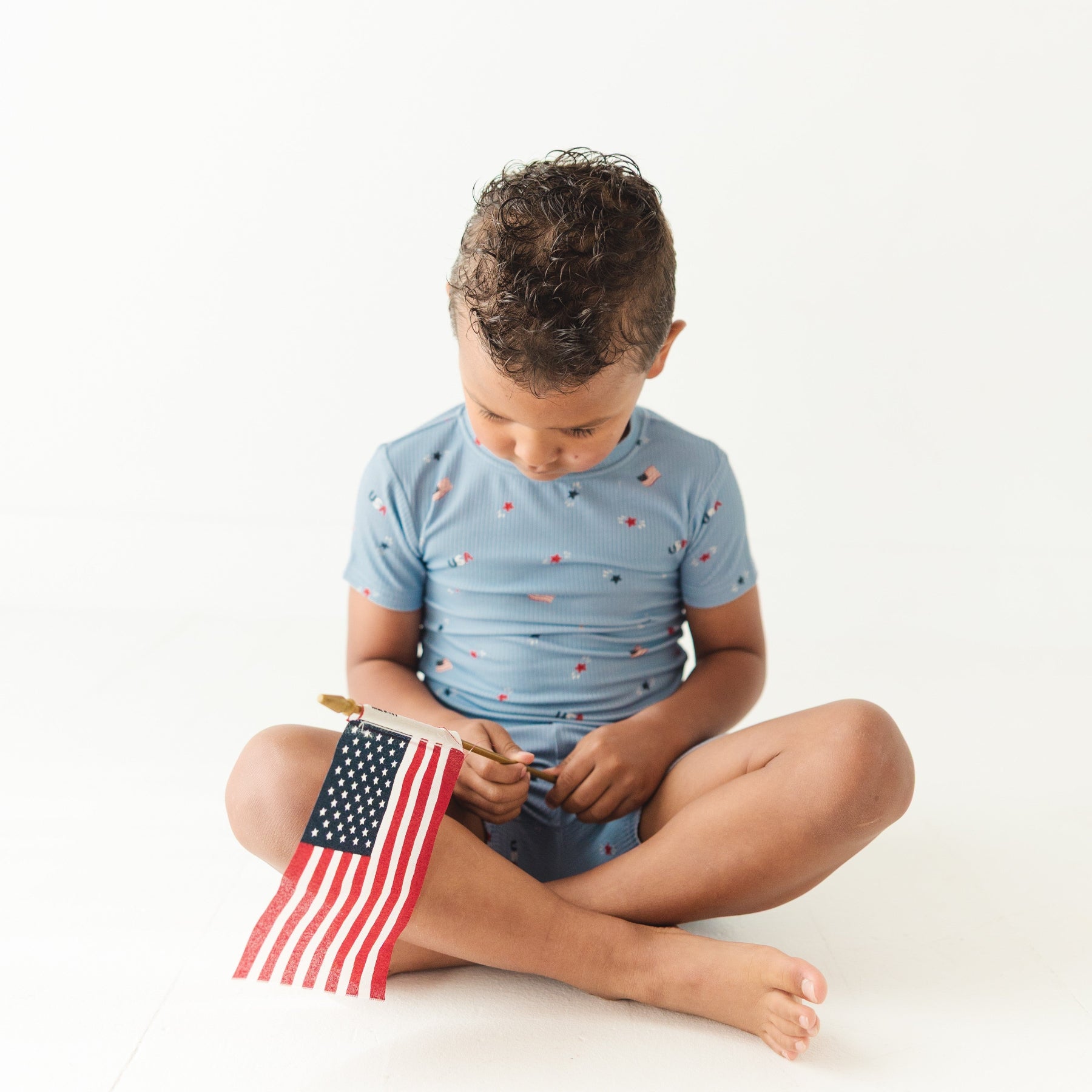 Child sitting on the floor wearing a light blue outfit with small American flag pattern, holding a miniature American flag.