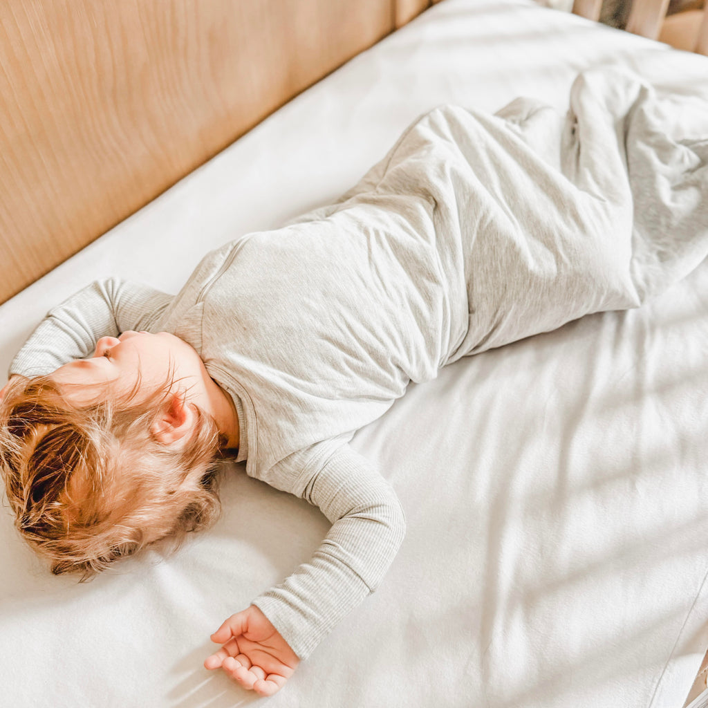 Child sleeping in a crib with white bedding, wearing a gray sleeping bag