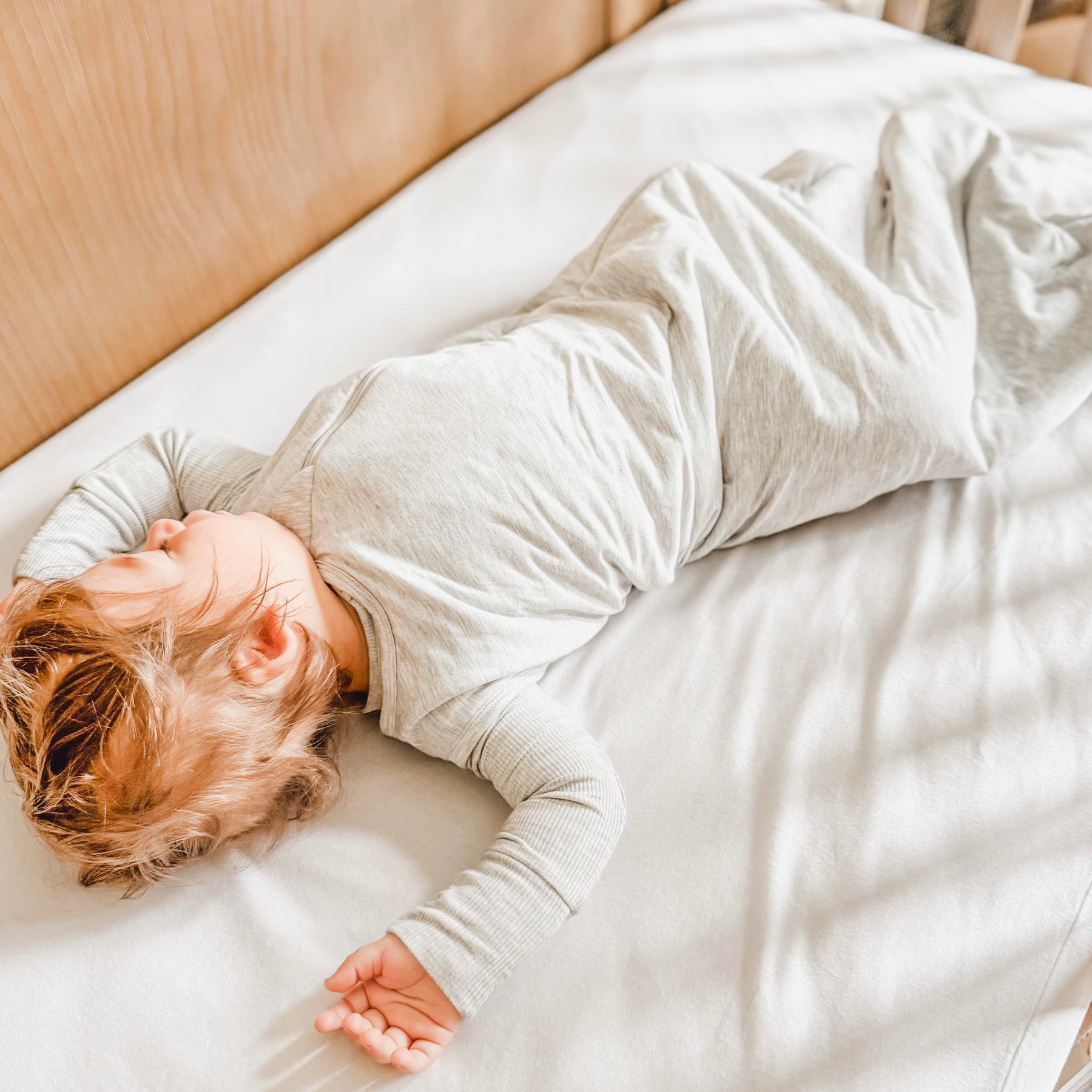 Child sleeping in a crib with white bedding, wearing a gray sleeping bag