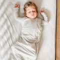 Child sleeping in a crib with white bedding, wearing a gray sleeping bag