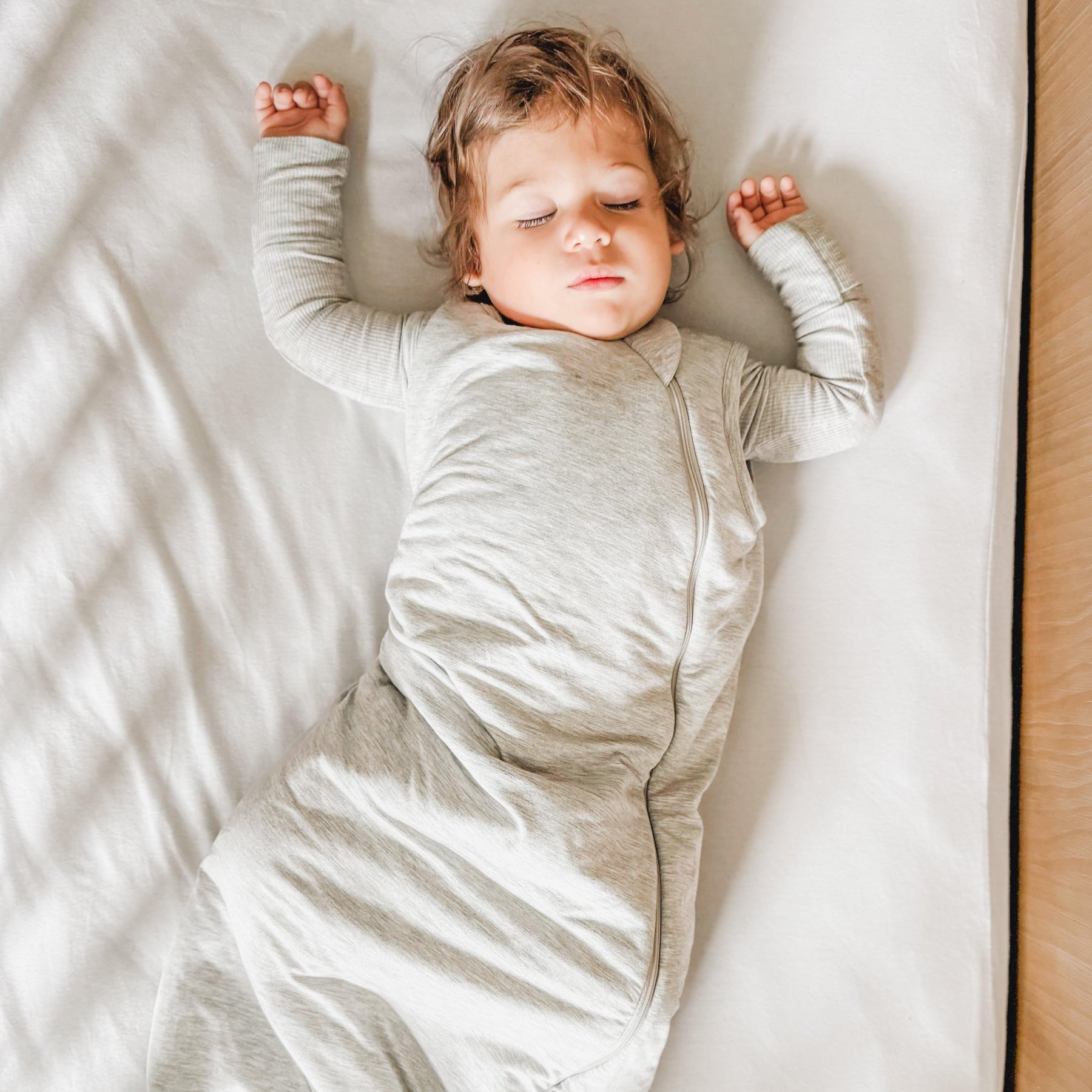 Child sleeping in a crib with white bedding, wearing a gray sleeping bag