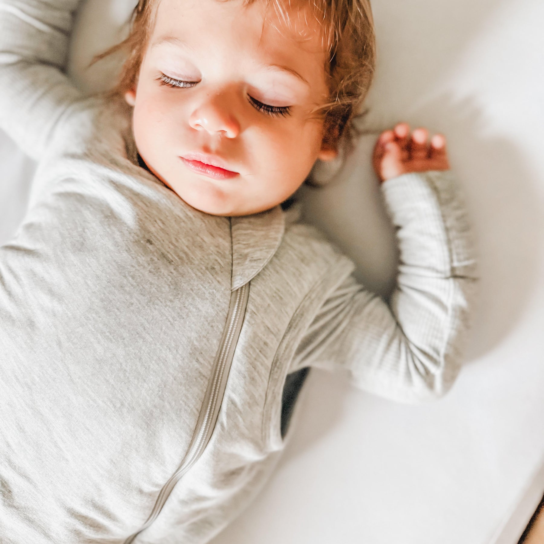 Close up of a child sleeping in a crib with white bedding, wearing a gray sleeping bag