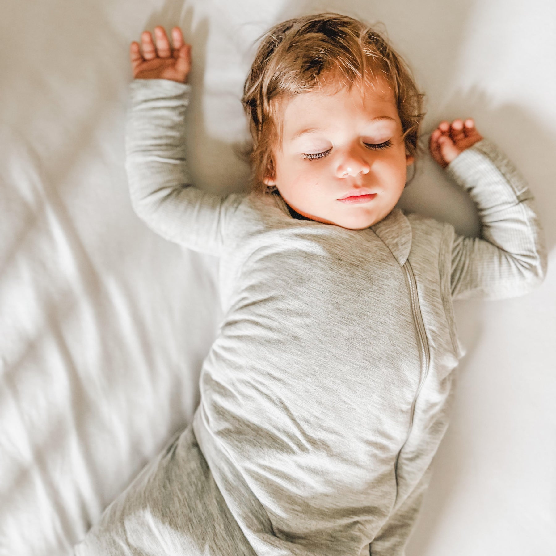 Child sleeping in a crib with white bedding, wearing a gray sleeping bag