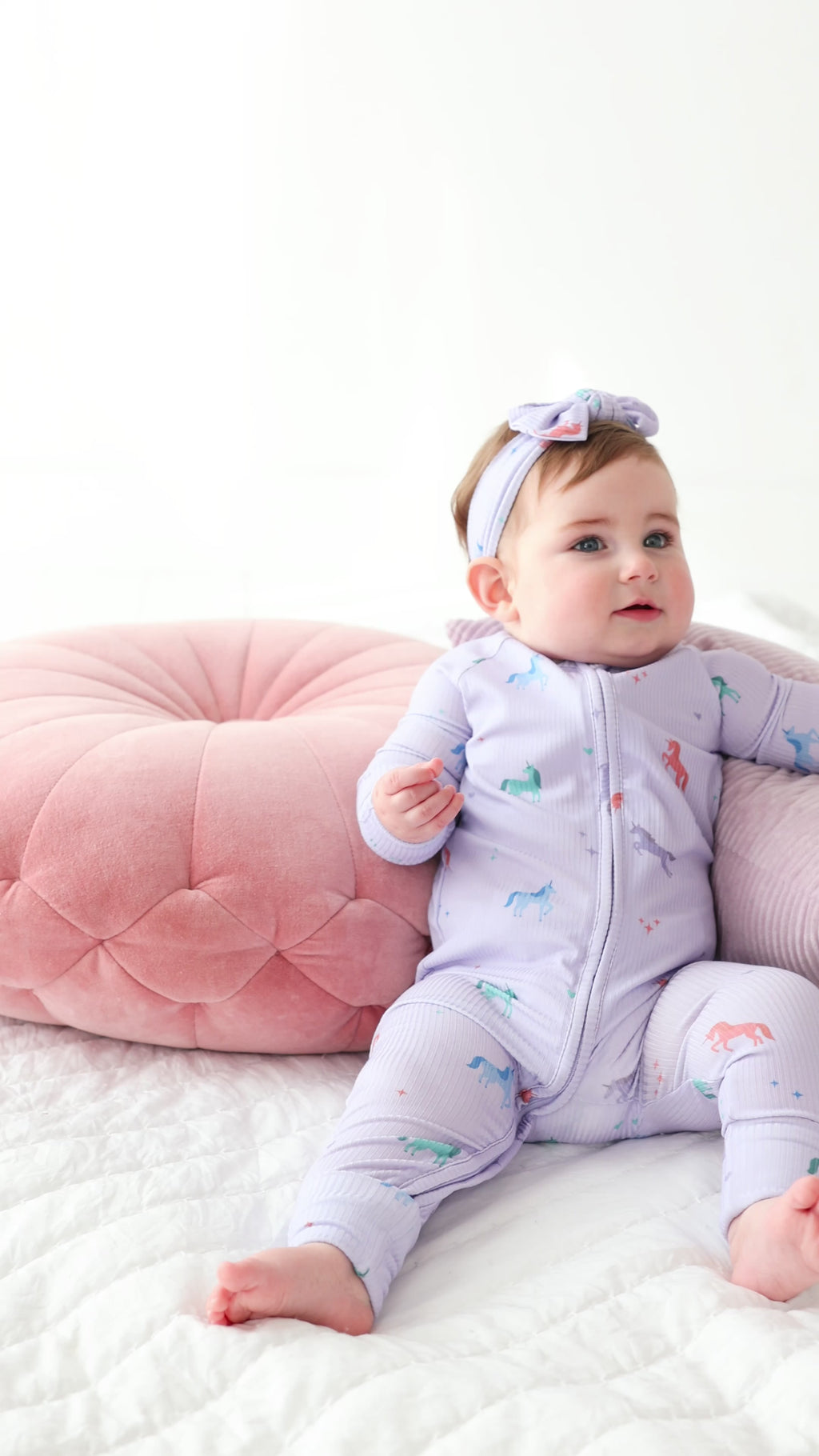 Video of a baby in a purple outfit with animal prints sitting on a white surface with pink and gray cushions.