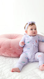 Video of a baby in a purple outfit with animal prints sitting on a white surface with pink and gray cushions.