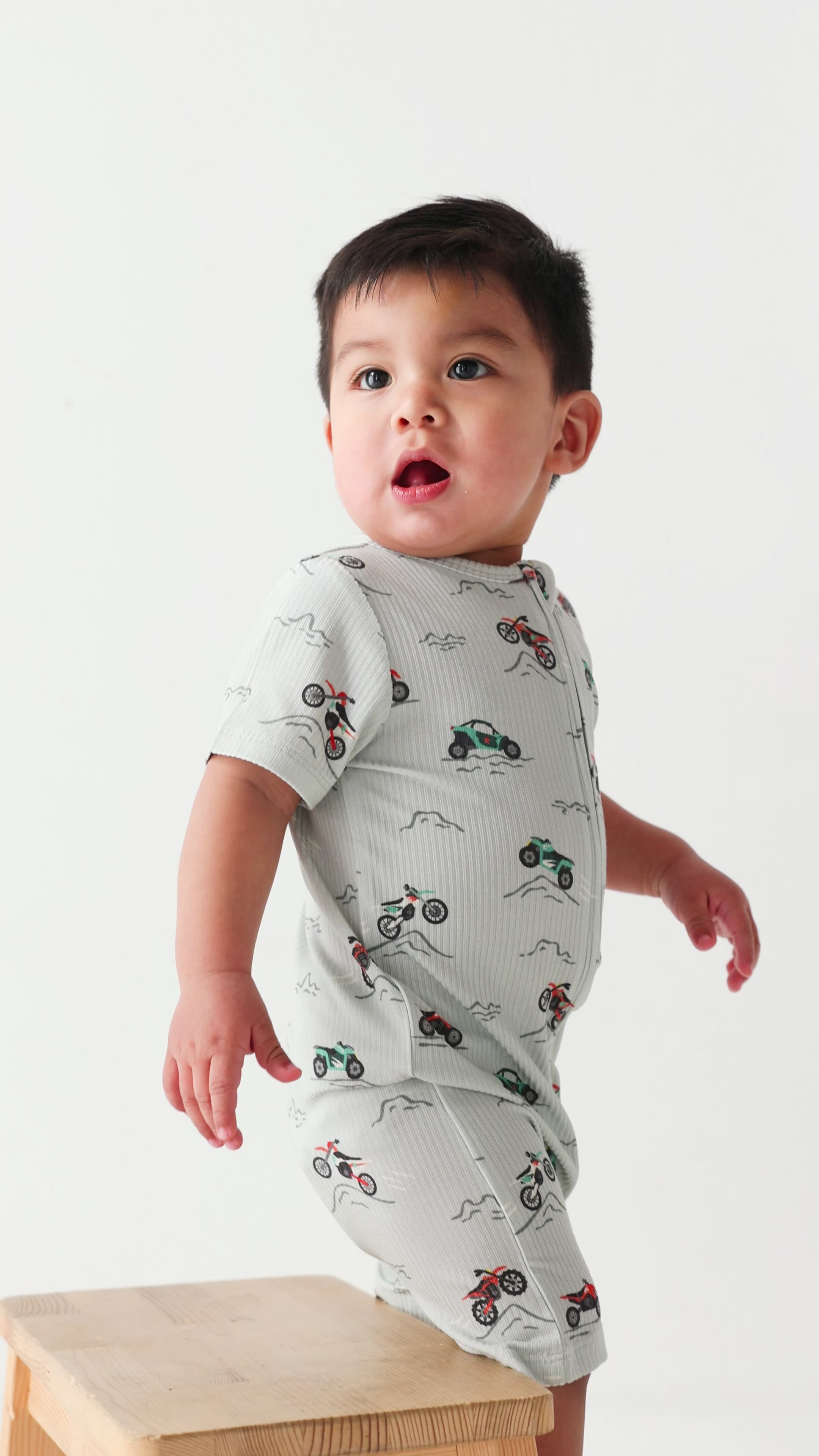Baby wearing a light gray onesie with vehicle patterns on a white background