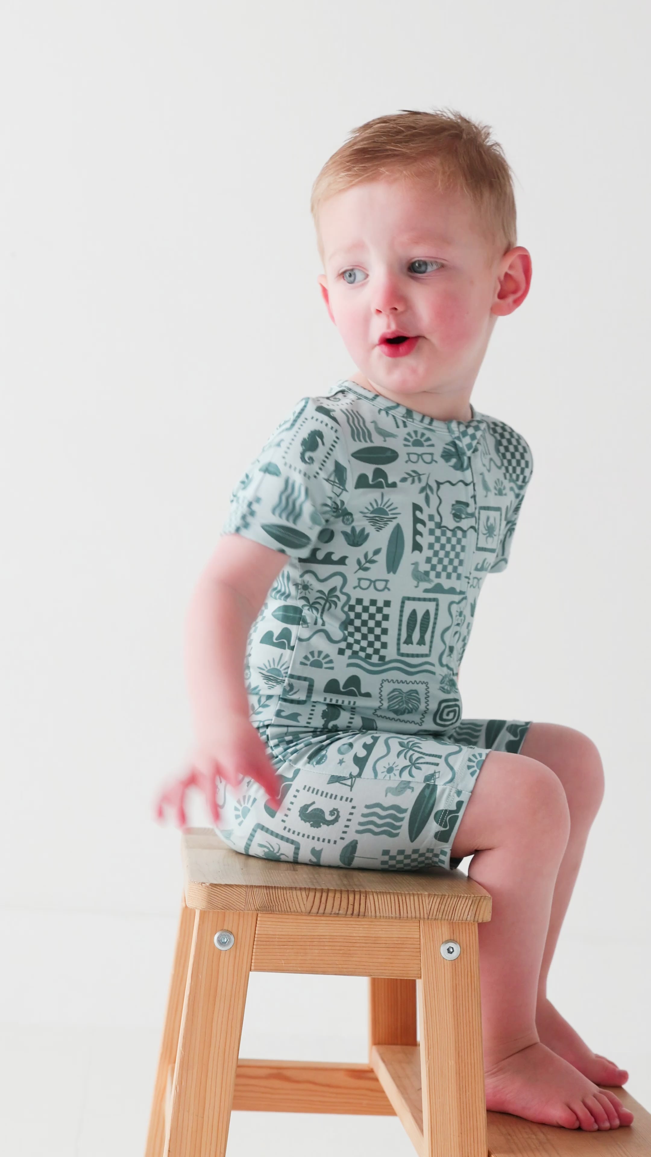 Child wearing a blue short romper with summer patterns sitting on a stool