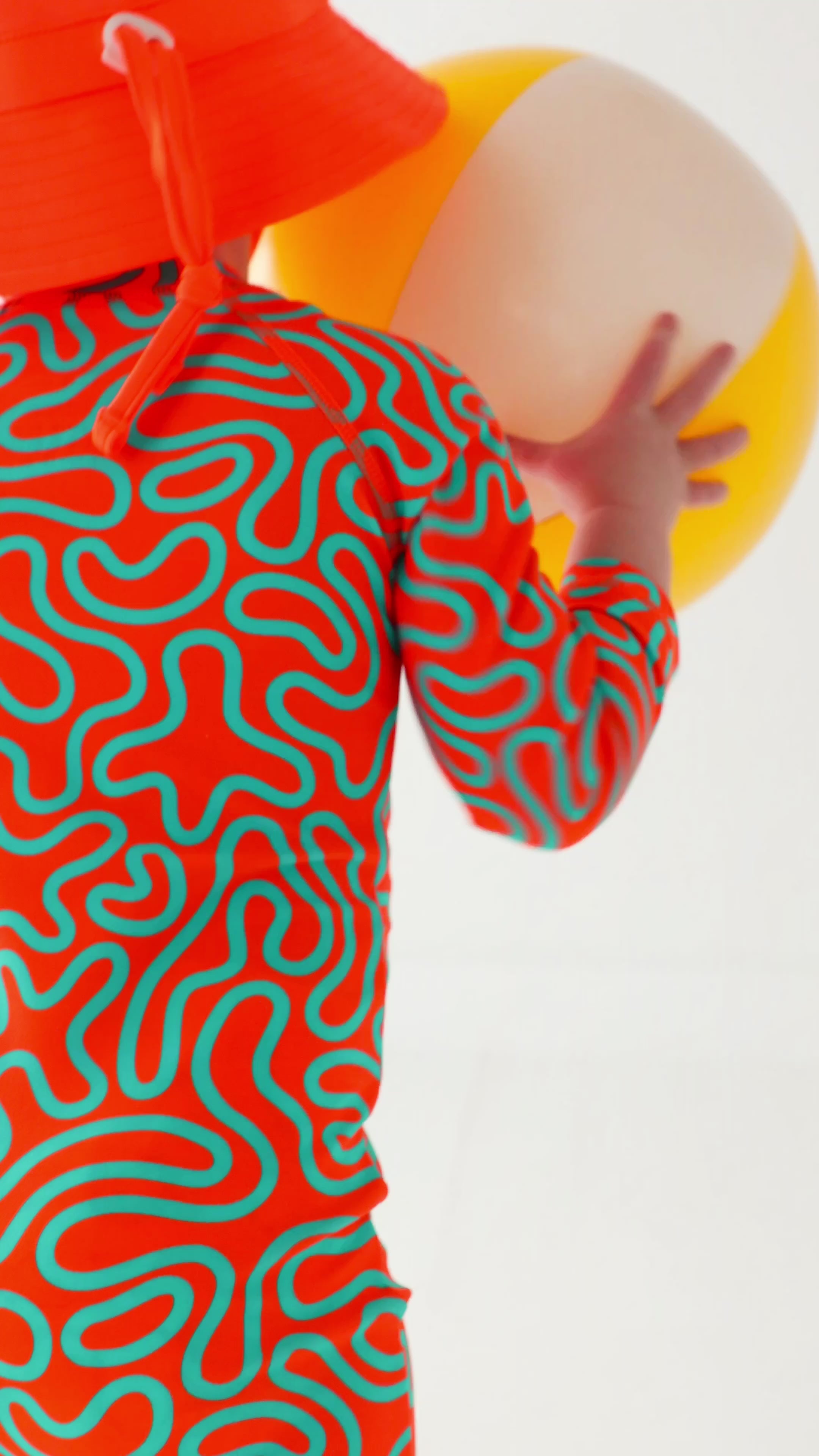 Child wearing a colorful swimsuit with a squiggly pattern and an orange bucket hat on a white background, playing with a white and yellow-striped beach ball