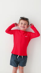 Child wearing a red rash guard, dark blue shorts and sunglasses on a white background