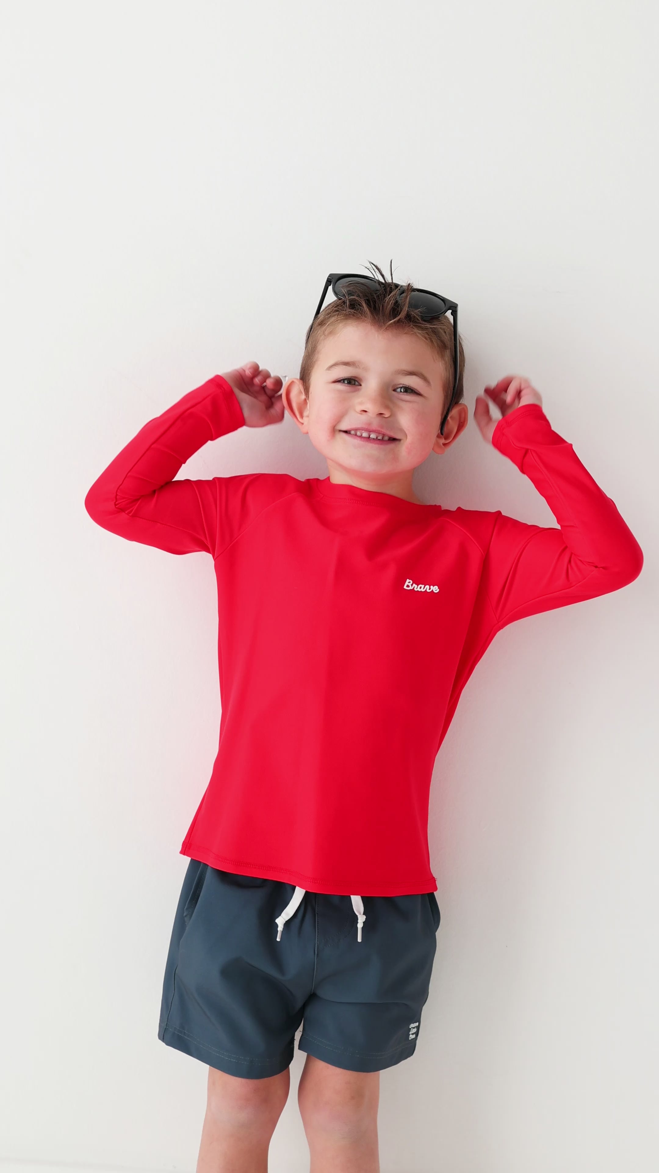 Child wearing a red rash guard, dark blue shorts and sunglasses on a white background