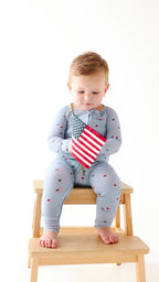 Video of a child wearing a light blue onesie with stars and flag patterns, holding a USA flag while sitting on a stool.