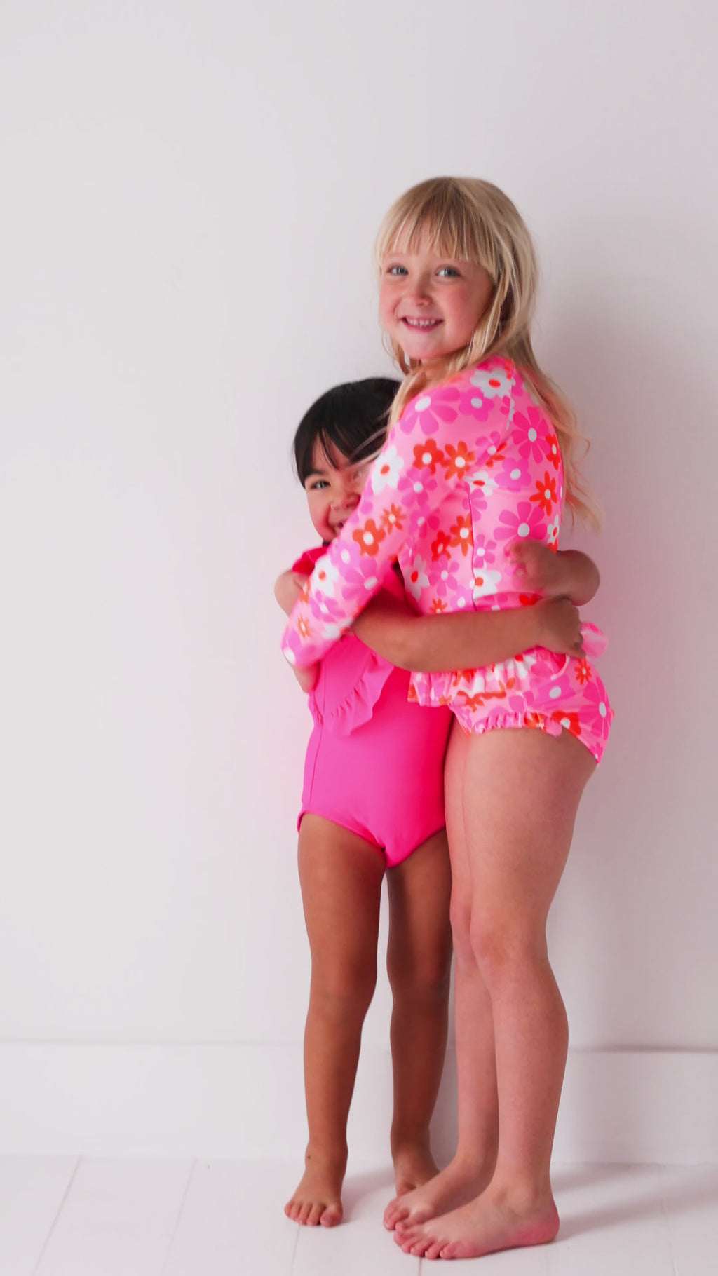 Two children in matching pink swimsuits standing on a white background