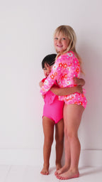 Two children in matching pink swimsuits standing on a white background