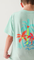 Back view of a child wearing a light blue t-shirt with colorful palm tree design on a white background