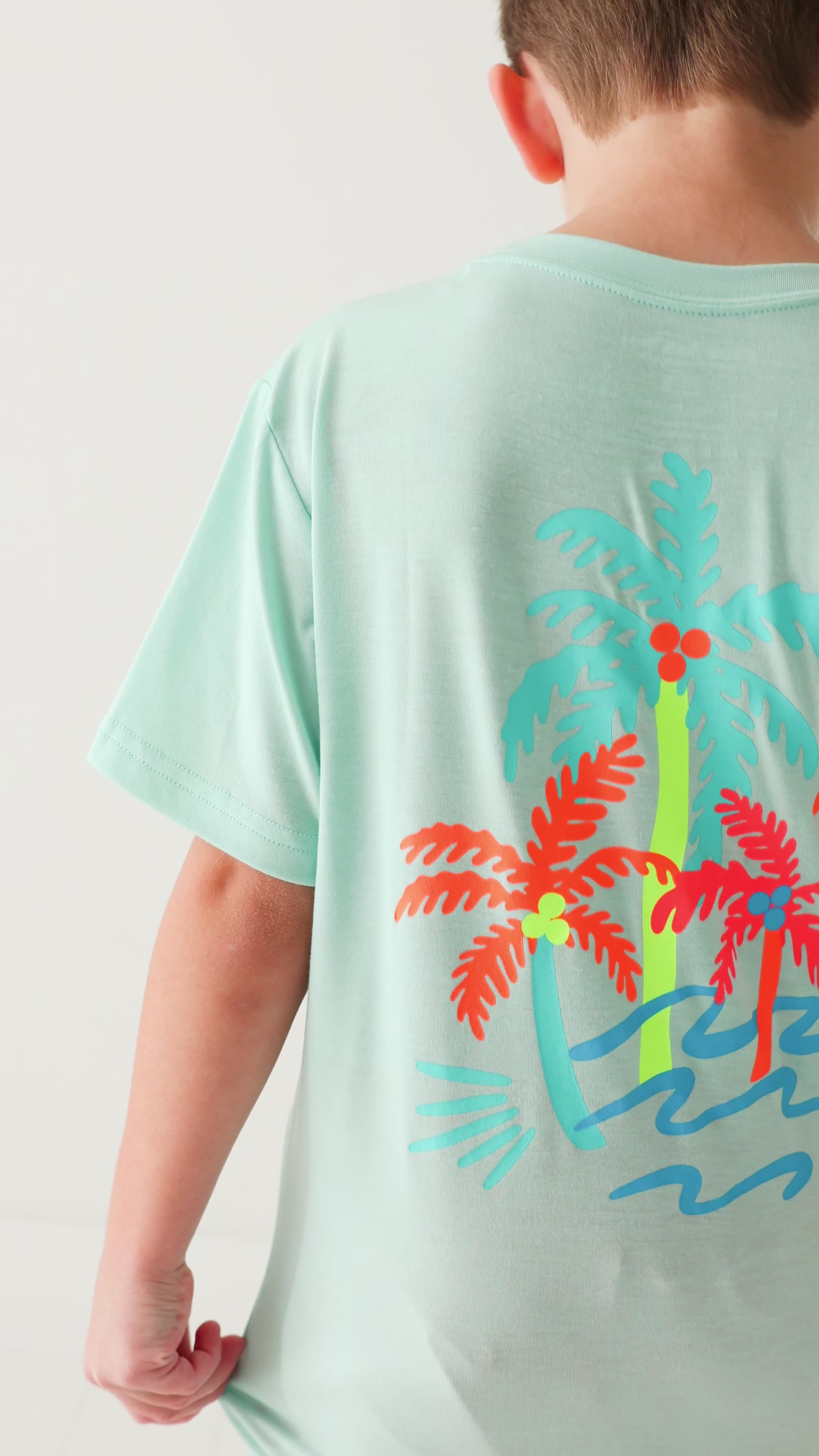 Back view of a child wearing a light blue t-shirt with colorful palm tree design on a white background