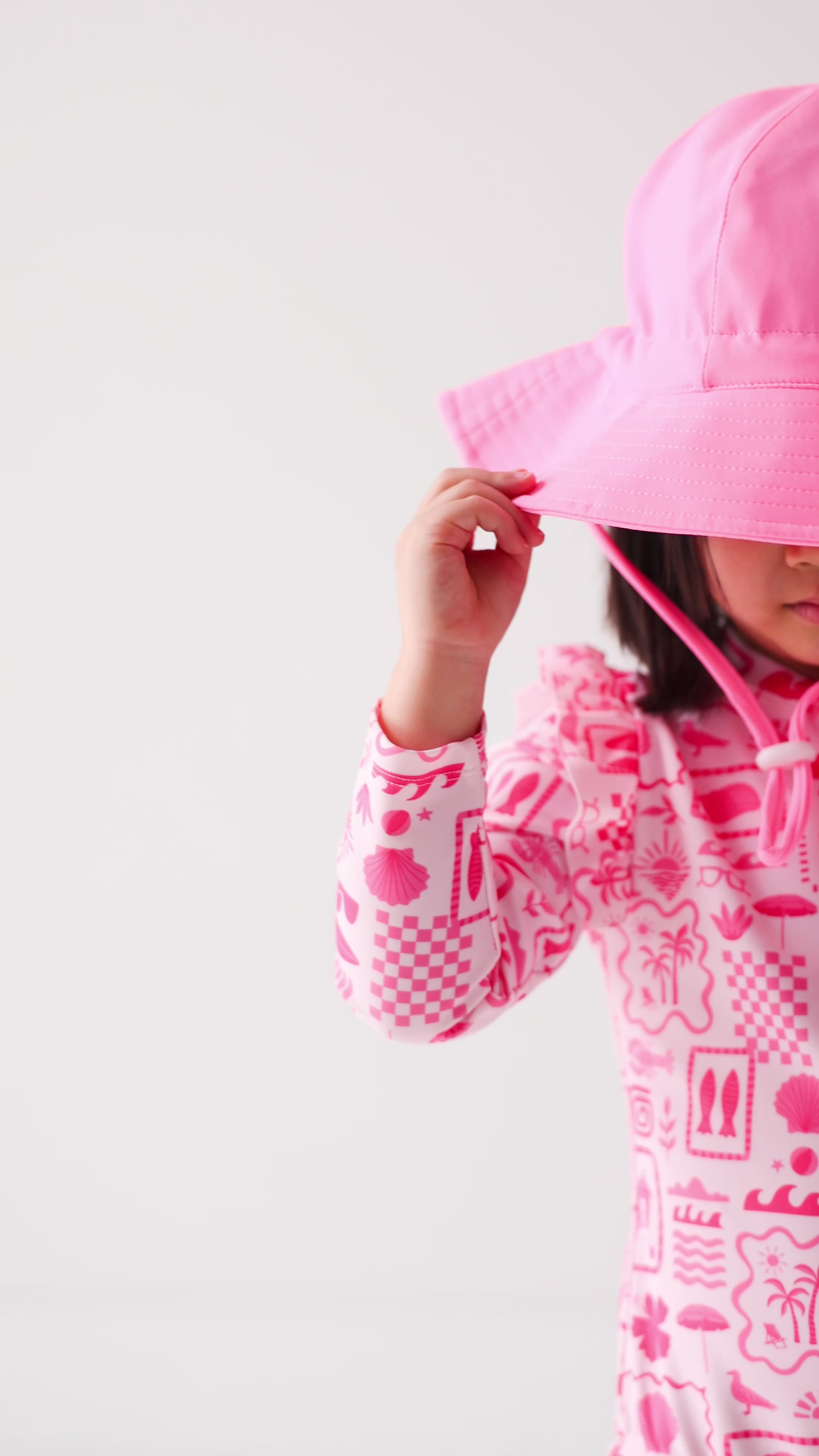 Child wearing a pink and white patterned long sleeve one-piece swimsuit and a matching light pink sun hat on a white background