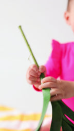 Baby wearing a bright pink one-piece swimsuit sitting on a white background with a yellow towel and green leaves.