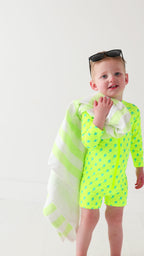 Child wearing a bright yellow boy swimsuit with a lightning bolt pattern and sunglasses on a white background, holding a green and white-striped towel