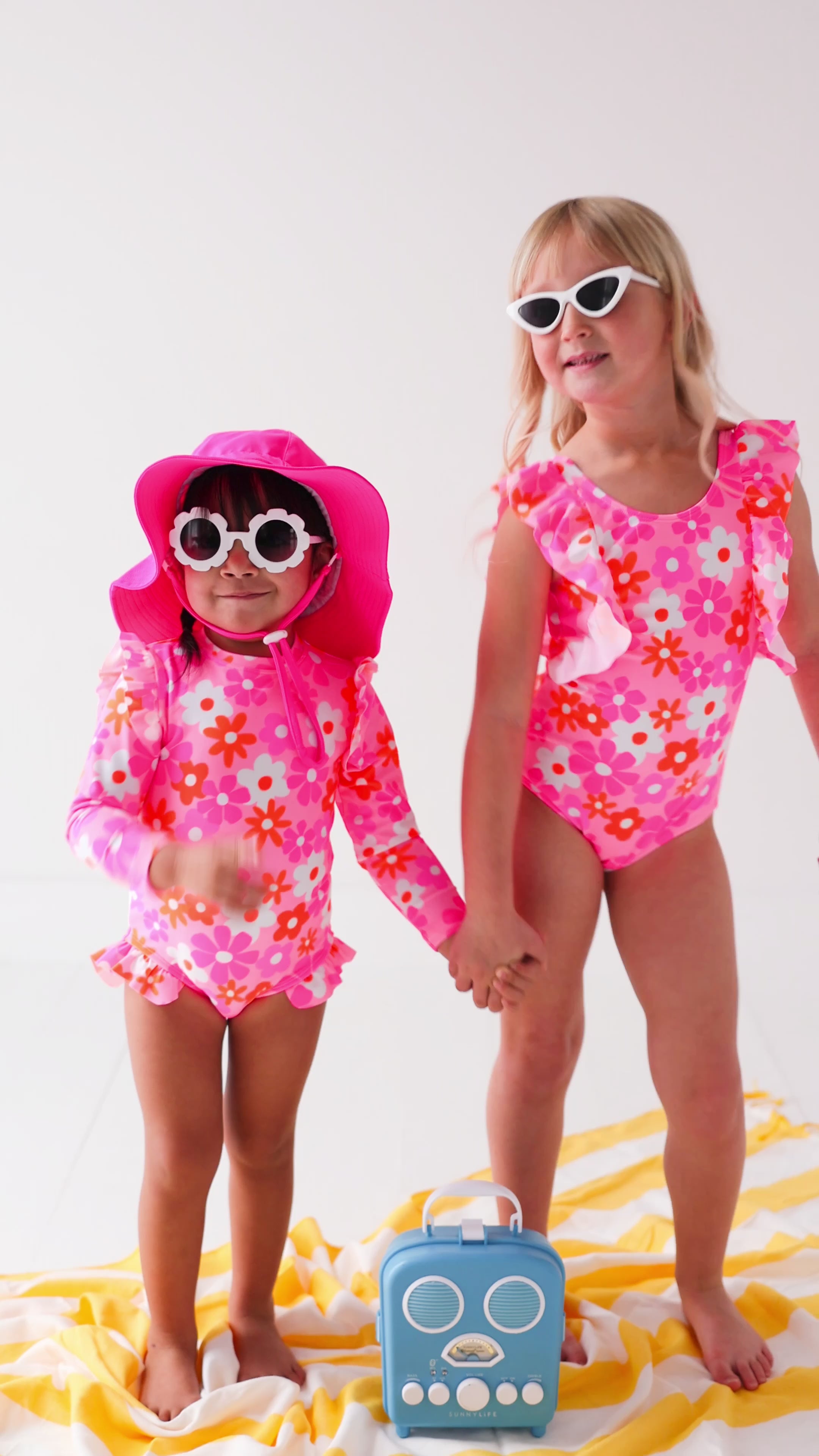 Two children in pink floral swimsuits sitting on a yellow and white striped towel with a blue radio toy.