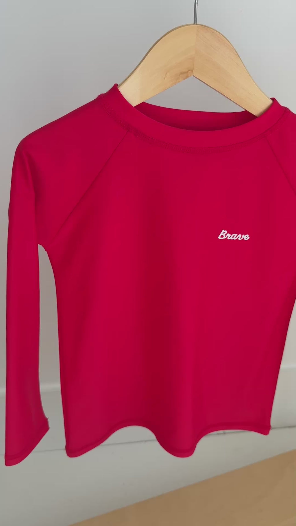 Red long-sleeve rash guard with 'Brave' logo hanging on a rack