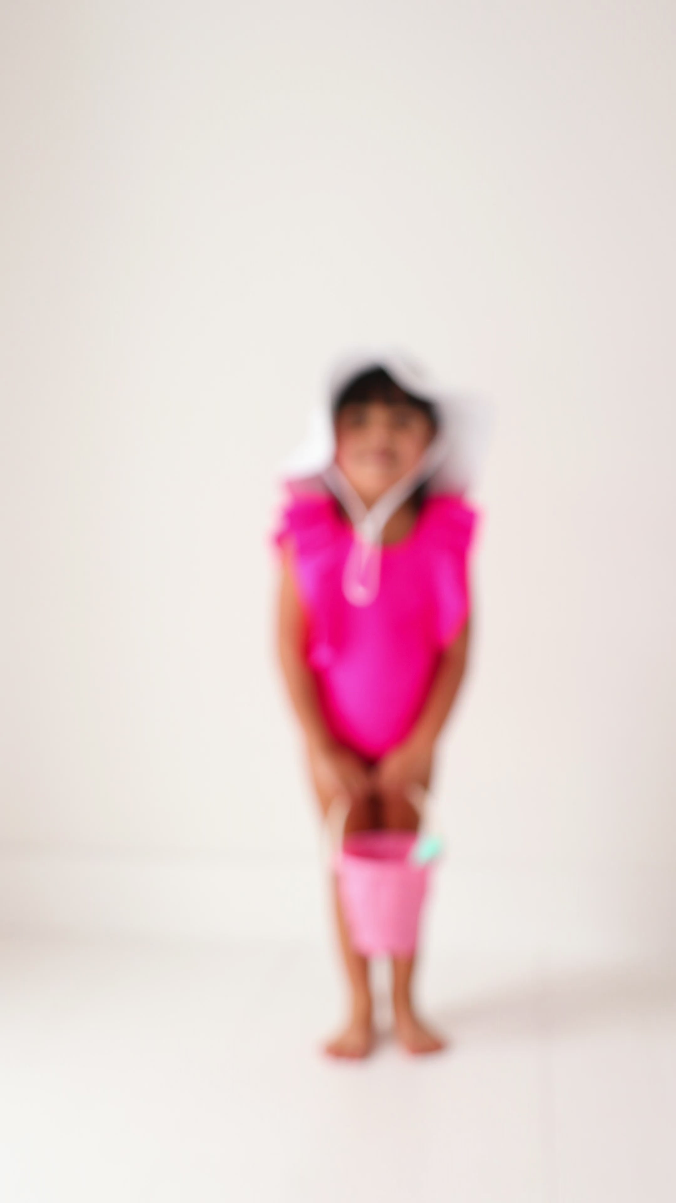A young child in a bright pink swimsuit and white hat playing with a sand bucket on a white background