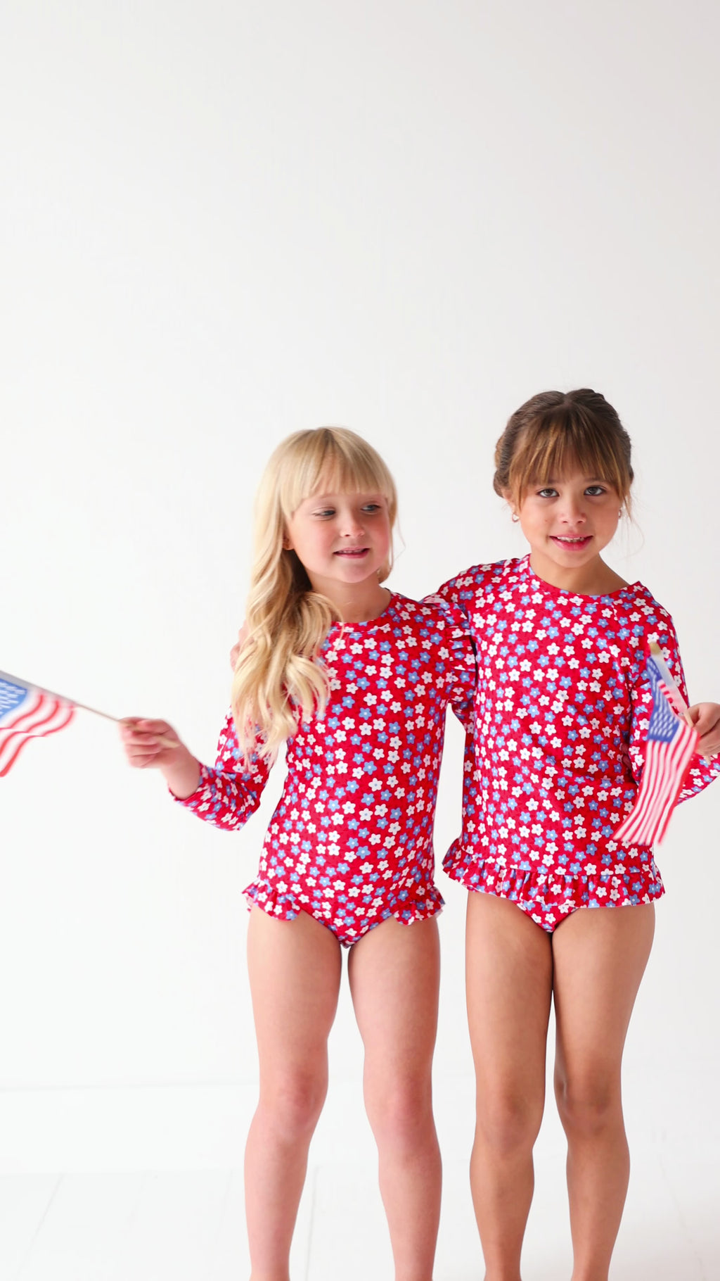 Two children wearing matching red and white floral patterned swimsuits, holding American flags, on a white background