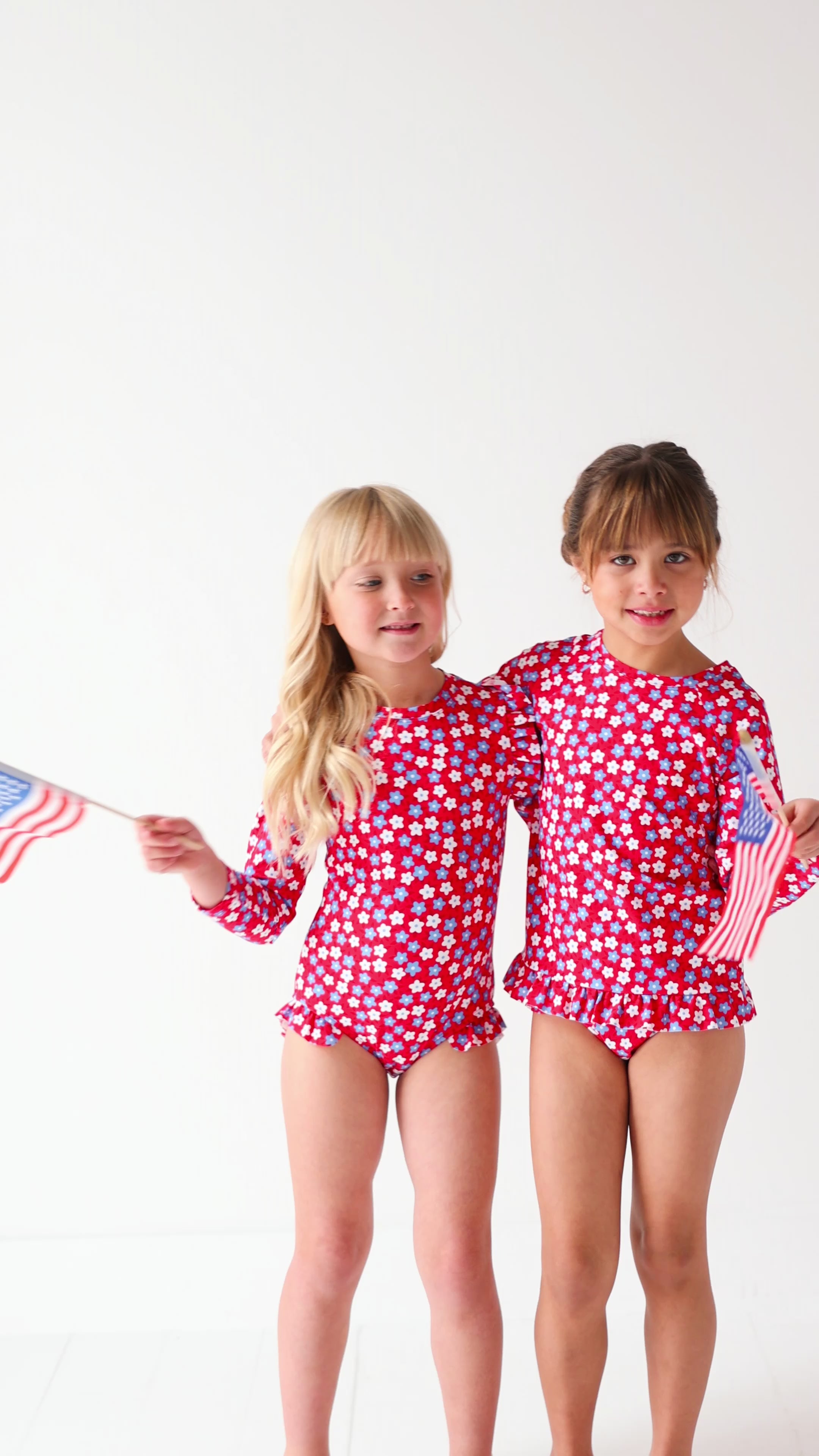 Two children wearing matching red and white floral patterned swimsuits, holding American flags, on a white background