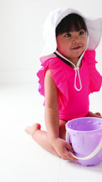 A young child in a bright pink swimsuit and white hat playing with a sand bucket on a white background