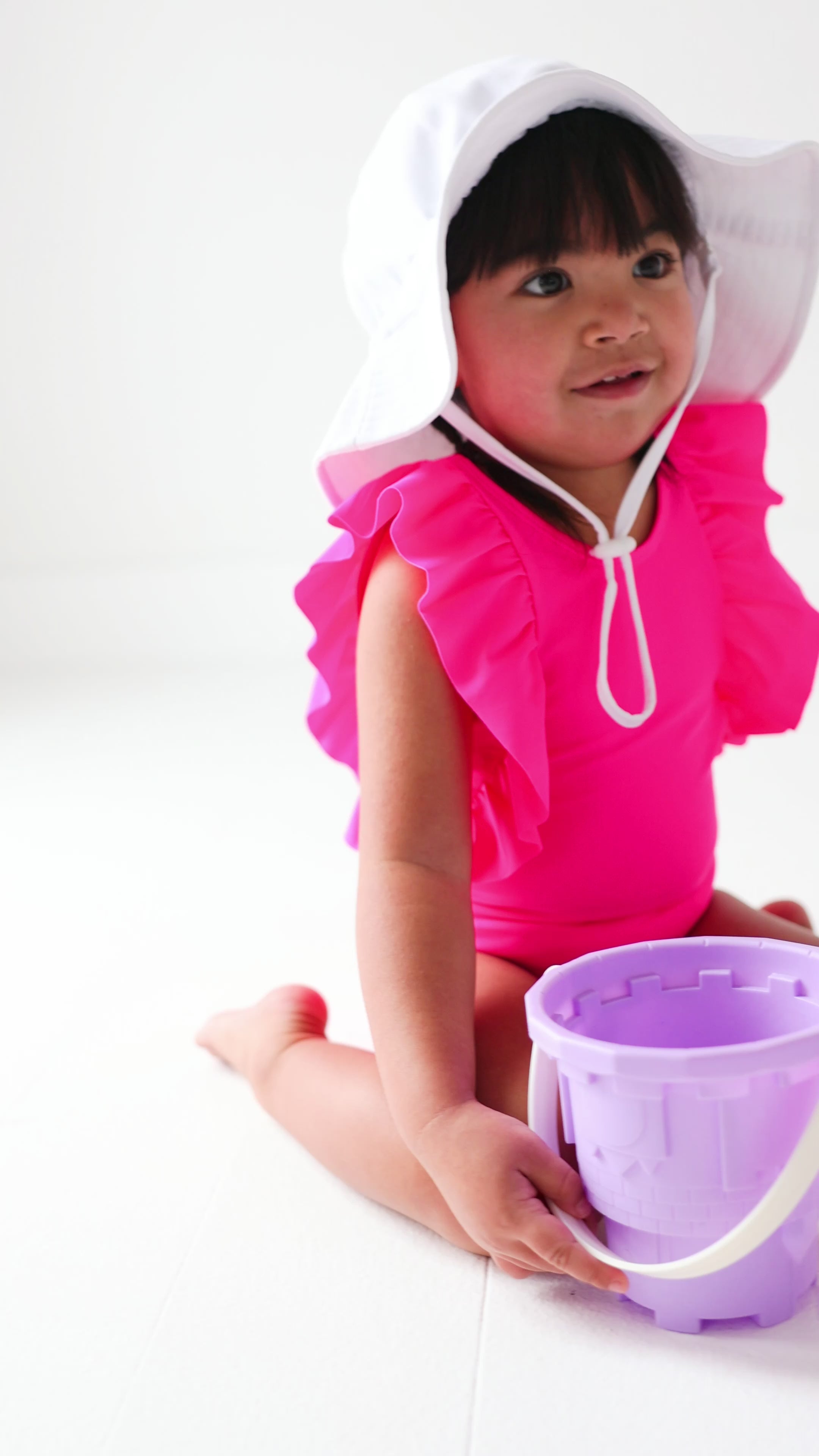 A young child in a bright pink swimsuit and white hat playing with a sand bucket on a white background