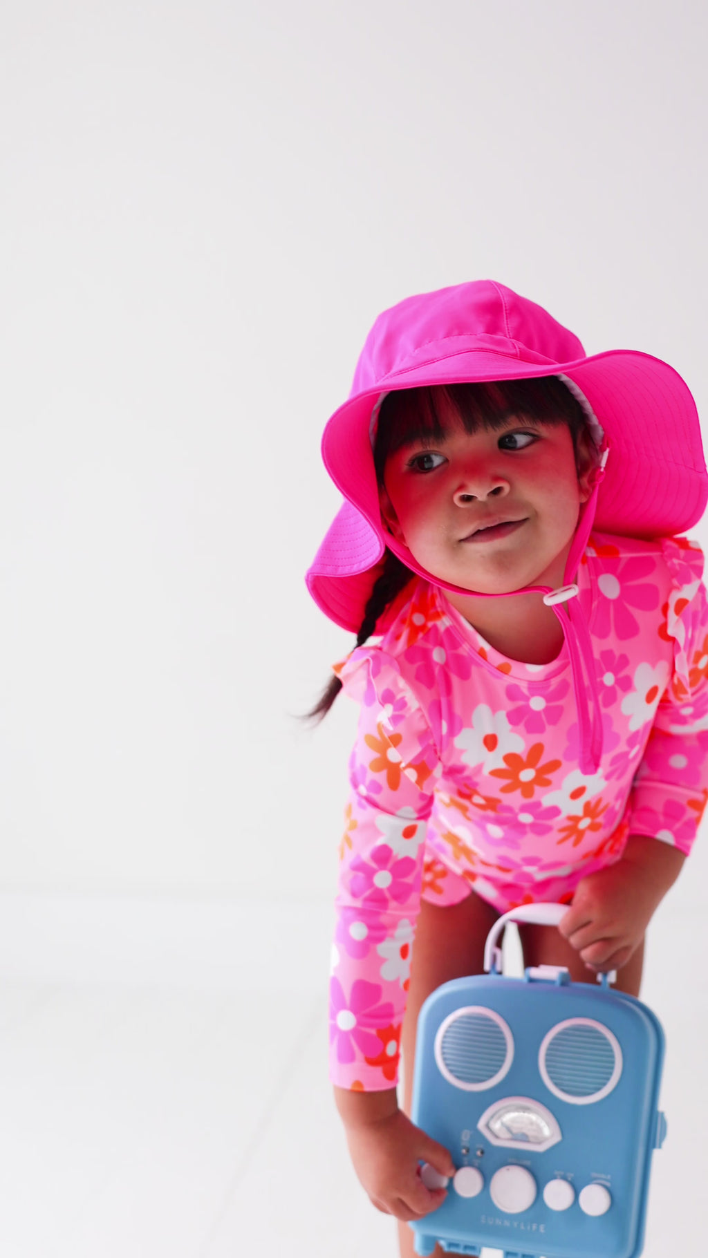 Child wearing a pink floral one-piece swimsuit and pink hat holding a blue radio toy on a white background