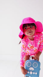 Child wearing a pink floral one-piece swimsuit and pink hat holding a blue radio toy on a white background