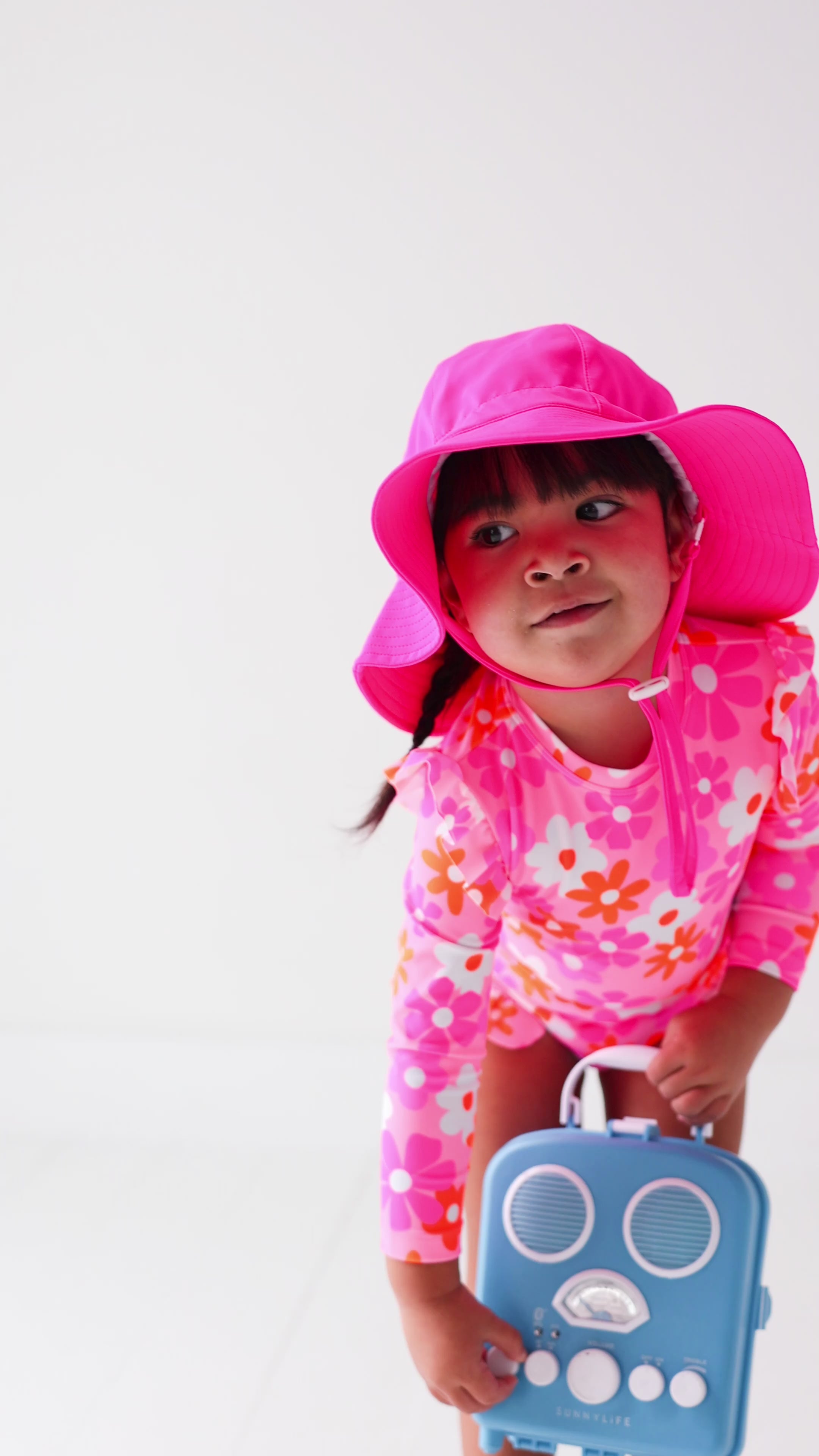 Child wearing a pink floral one-piece swimsuit and pink hat holding a blue radio toy on a white background