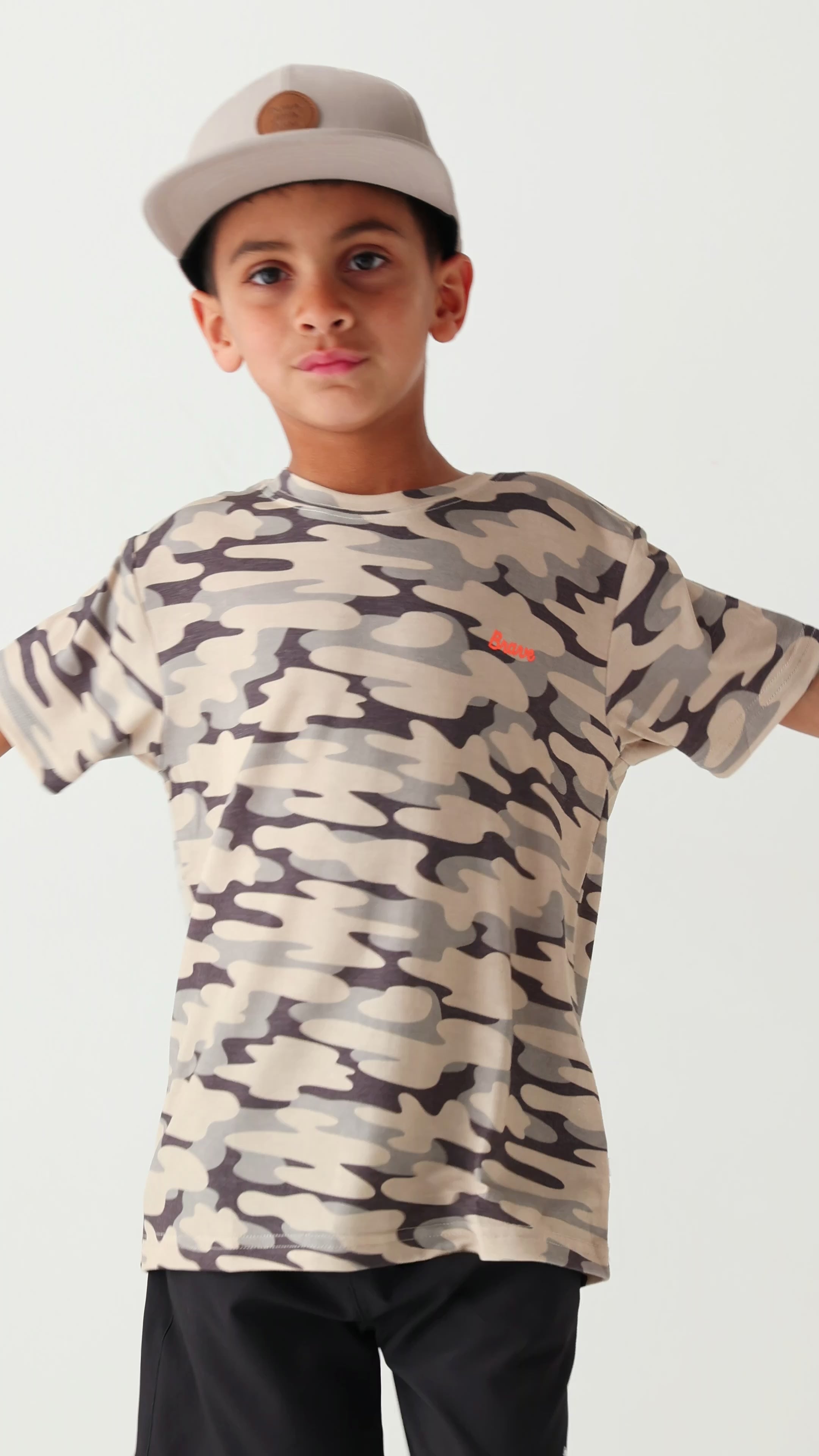 Child wearing a camouflage t-shirt, taupe cap and black shorts on a white background