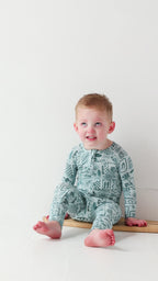 Child wearing a blue summer-patterned onesie sitting on a tiny surfboard on a white background
