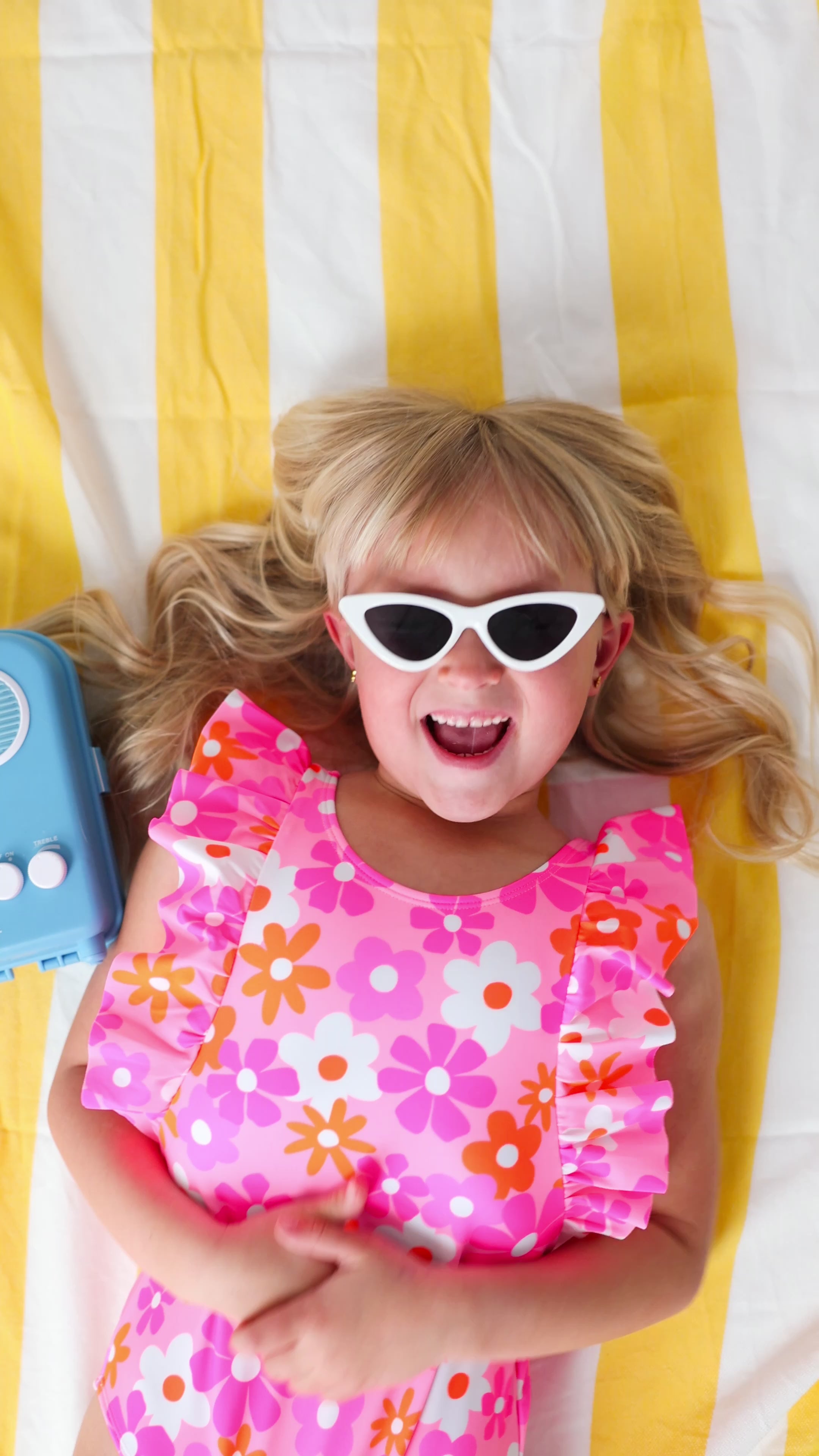 Child in a pink floral swimsuit with ruffles, wearing sunglasses, lying on a yellow and white striped fabric with a blue radio toy