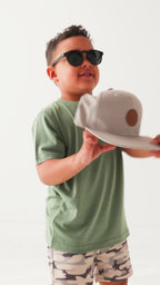 Child wearing a green t-shirt, taupe cap and camouflage shorts on a white background