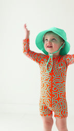 Child wearing a colorful swimsuit with a squiggly pattern and a turquoise bucket hat on a white background