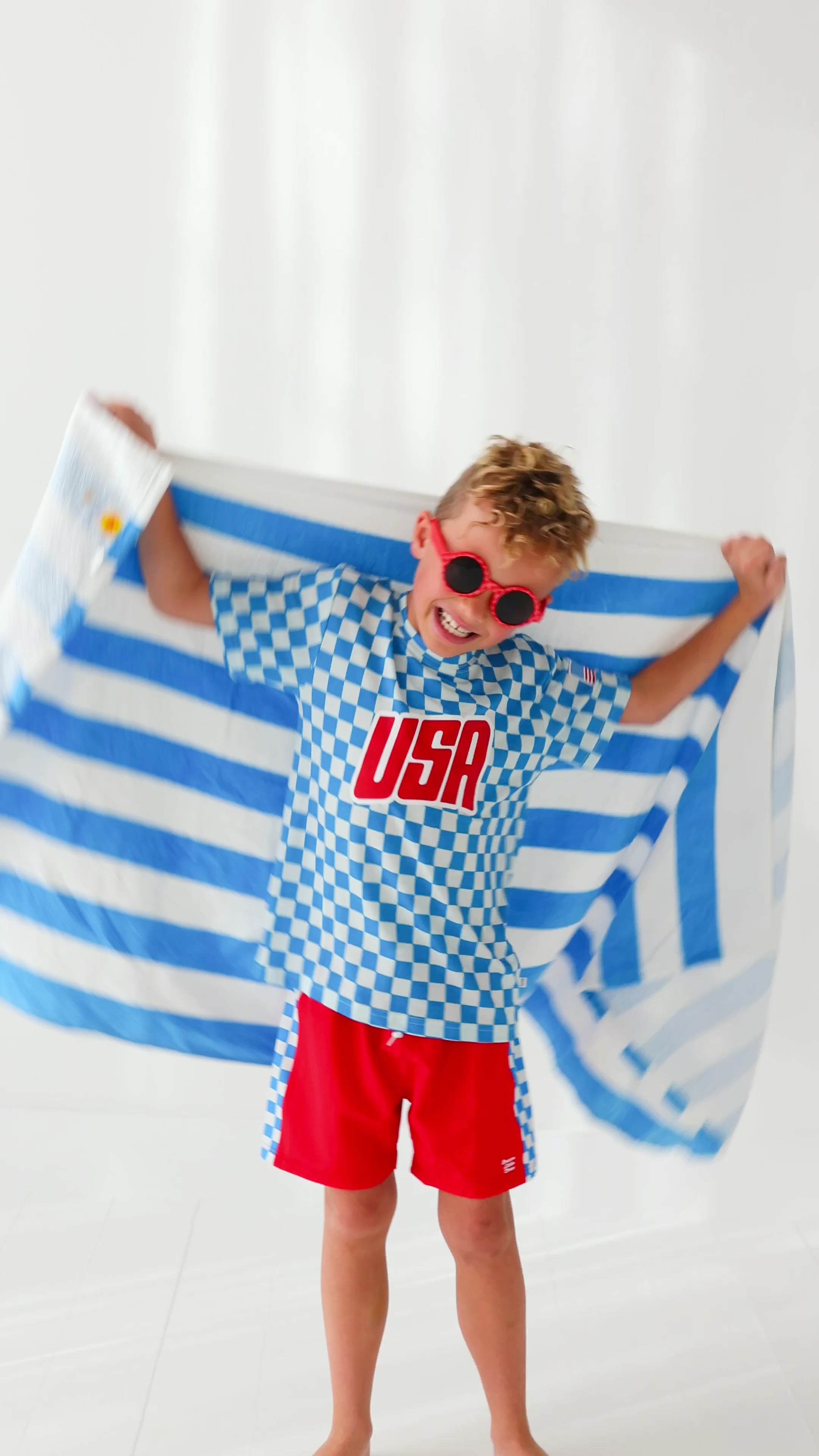 Child wearing a blue and white checkered shirt with 'USA' printed on it, standing against a blue and white striped towel fabric.