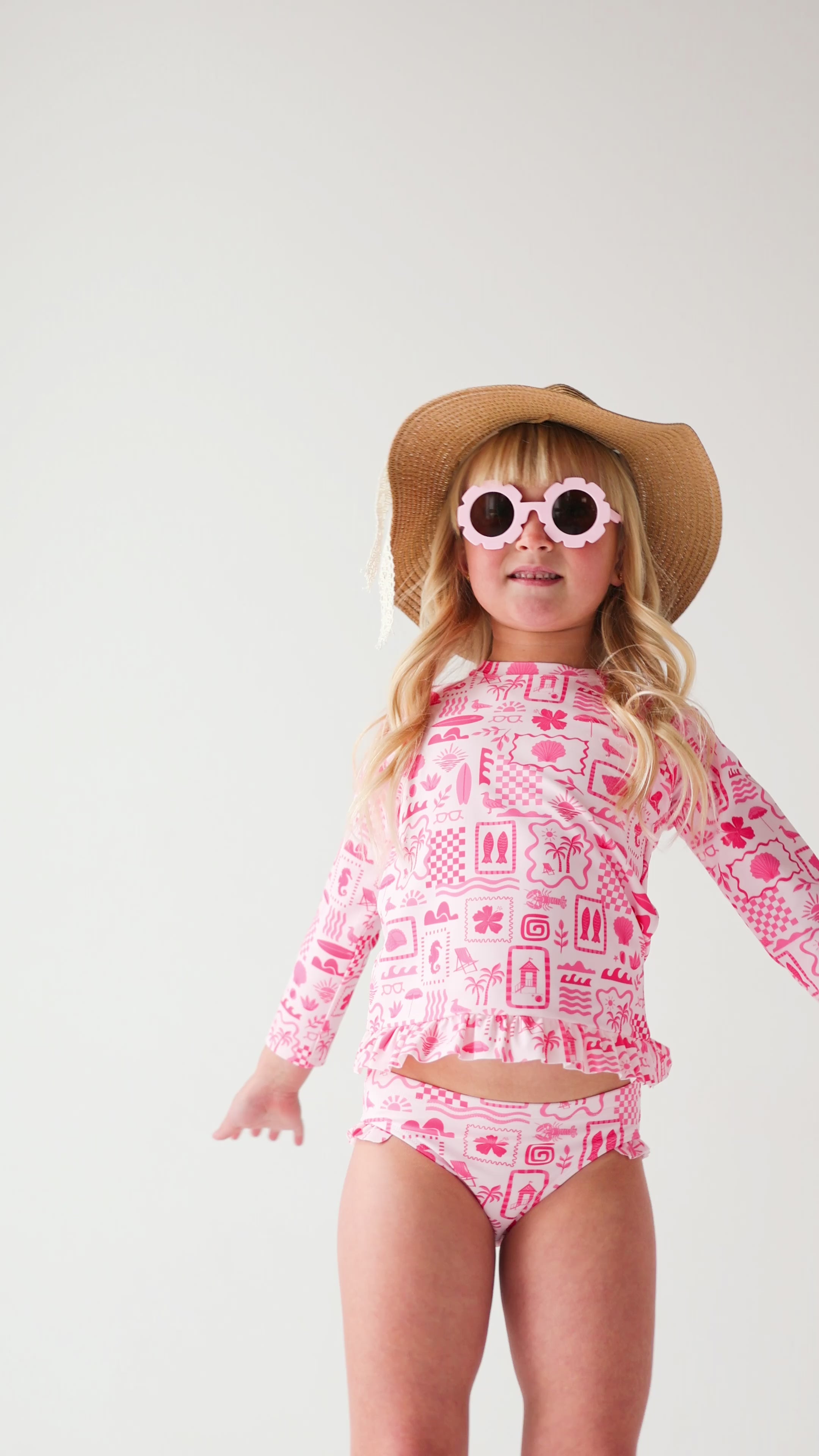 Child wearing a pink and white patterned long sleeve two-piece swimsuit, floral sunglasses and brown hat on a white background