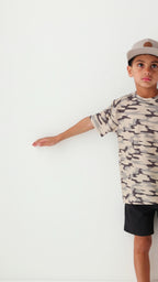 Child wearing a camouflage t-shirt, taupe cap and black shorts on a white background