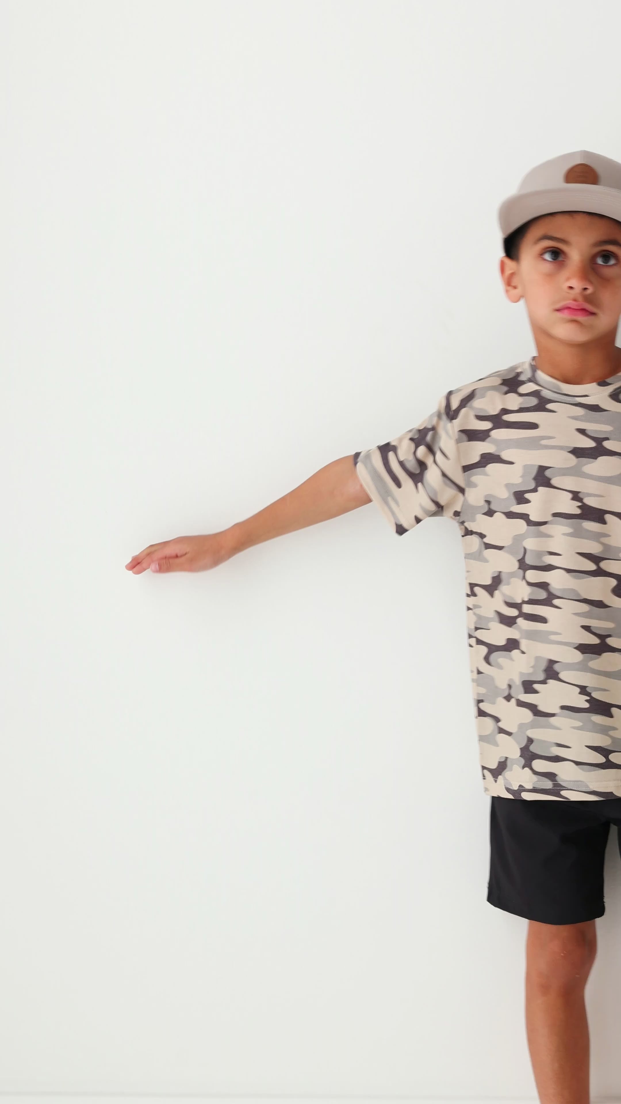 Child wearing a camouflage t-shirt, taupe cap and black shorts on a white background